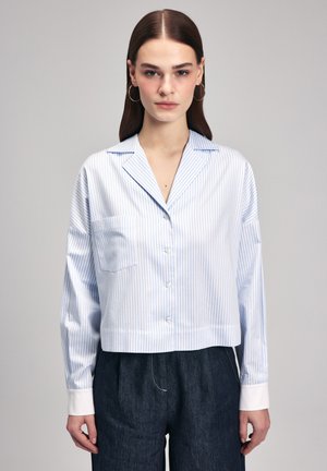 Camicia a righe in blu chiaro e bianco, maniche lunghe, design cropped, chiusura a bottoni, taschino sul petto, texture liscia, abbinata a pantaloni in denim scuri.