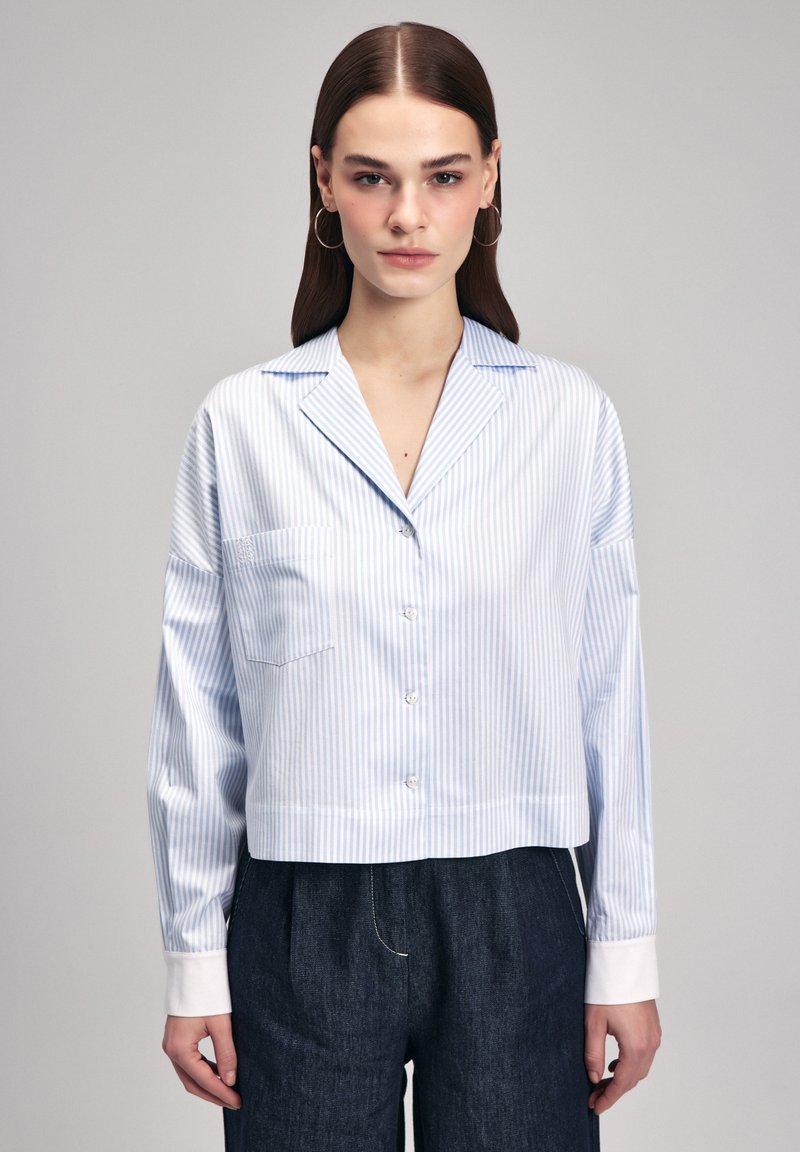 Camicia a righe in blu chiaro e bianco, maniche lunghe, design cropped, chiusura a bottoni, taschino sul petto, texture liscia, abbinata a pantaloni in denim scuri.