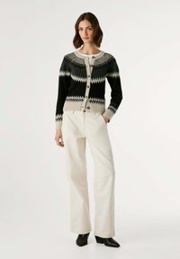 Cardigan à motifs noir et crème avec des boutons, encolure ronde et poignets côtelés. Associé à un pantalon droit blanc et des bottines noires.