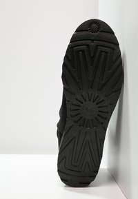 Suela de zapato de goma negra con un patrón texturizado en zigzag y un emblema. El ángulo muestra la parte inferior con un dibujo bien definido y un diseño circular.