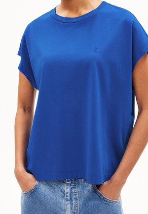 Osoba ubrana w luźny, jasnoniebieski top z krótkim rękawem, lekko wpuszczony w jasnoniebieskie dżinsy, z rękami w kieszeniach.