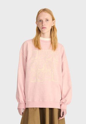 Jeune femme aux longs cheveux roux portant un sweat-shirt rose ample avec un imprimé graphique discret et une jupe marron, debout devant un fond uni.