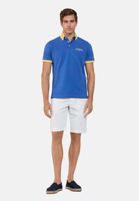 Niebieska koszulka polo z żółtymi akcentami kołnierzyka i logo, w połączeniu z białymi szortami i granatowymi espadrylami. Gładki materiał, luźny krój.