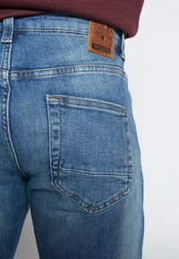 Lichtblauwe spijkerbroek met een zachte textuur, voorzien van een bruin leren label, stiksel op de achterzakken en een subtiel vervaagd ontwerp.