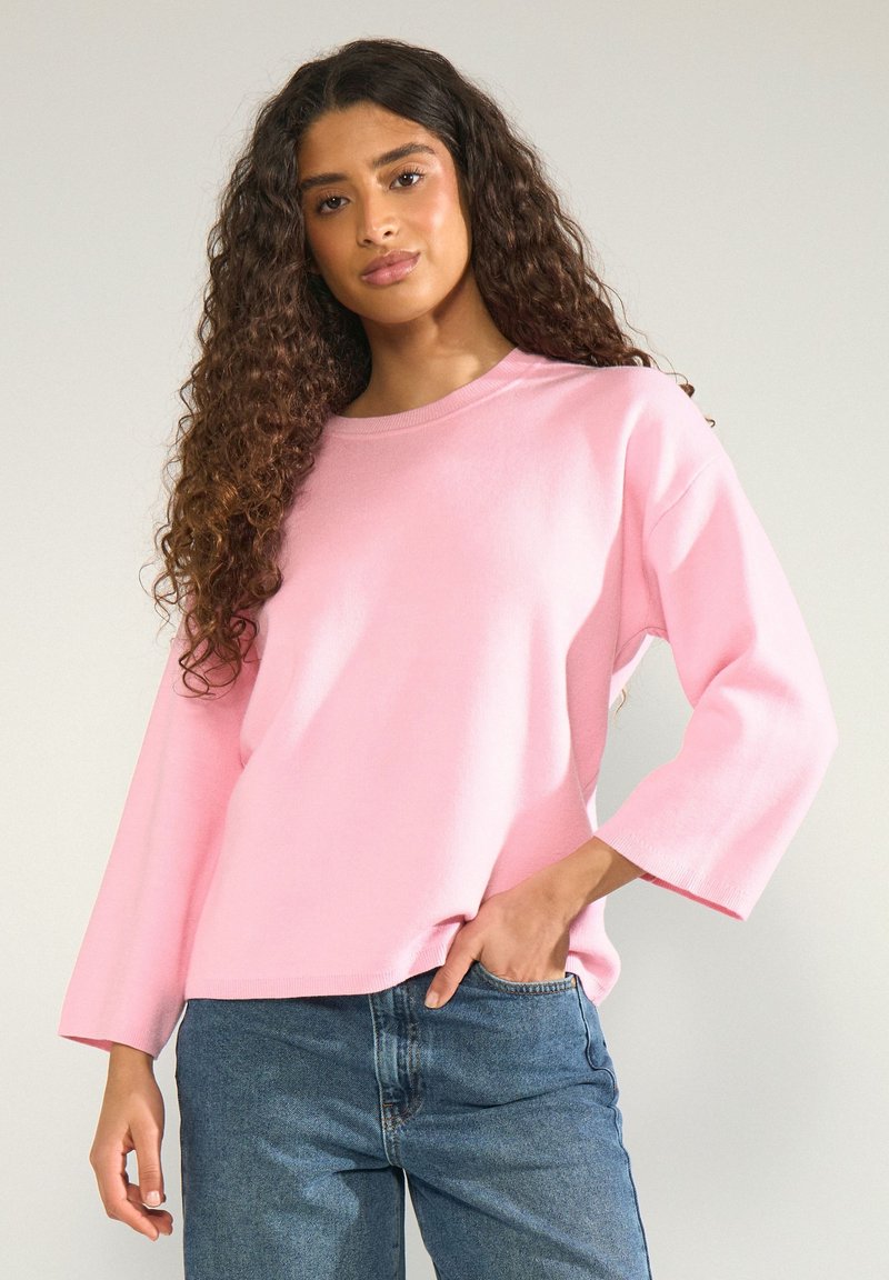 Femme aux longs cheveux bouclés portant un haut rose ample à manches longues et un jean bleu, debout avec une main dans la poche, sur fond uni.