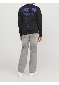 Svart sweatshirt med blå grafisk text på ryggen, kombinerad med ljusgrå jeans och vita sneakers. Enkel, avslappnad design.
