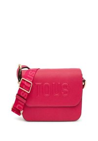 Bolso cruzado rosa con logo "TOUS" en relieve, material texturizado, herrajes dorados y una correa ajustable rosa en contraste con branding repetido.