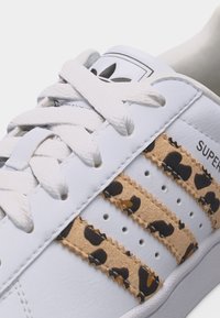 Baskets blanches avec des lacets blancs, présentant trois bandes beiges ornées d'un imprimé léopard noir et marron, et le logo Adidas sur la languette.