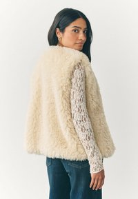 Kreemikollane faux karusnahast vest, millel on pehme, fluffy tekstuur. Omab lõdva istuvust ilma kinnitusteta, kantakse pitsilisest pikkade varrukatega pluusi peal.