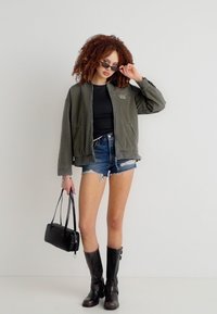 Γυναίκα με σγουρά μαλλιά που φοράει γυαλιά ηλίου, πράσινο bomber μπουφάν, μαύρο τοπ, τζιν σορτς, μαύρες μπότες και κρατάει μια μαύρη τσάντα χειρός.