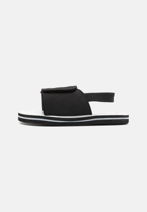 Sandale noire avec un upper texturé, comportant une seule sangle ajustable et une semelle blanche. Rayuresnoires et blanches contrastées sur la semelle extérieure.