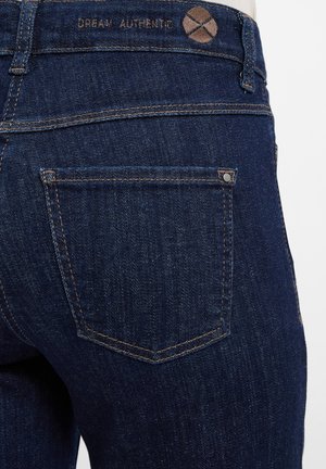 Donkerblauwe denim jeans met een gestructureerd oppervlak, voorzien van een rechthoekige achterzak, minimalistische stiksels en een label met de opdruk "DREAM AUTHENTIC."
