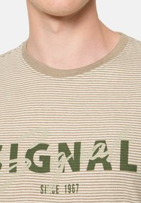 Camiseta de rayas beige y crema con escote redondeado, que presenta el texto verde "SIGNAL" y un logo impreso con efecto polvo, fabricada en suave material de algodón.
