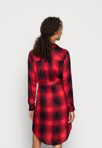 Rochie cambrată cu carouri roșii și negre, cu mâneci lungi, margine rotundă și curea în talie. Fabricată dintr-un material ușor, cu o textură netedă.