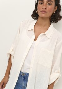 Chemise en lin blanche avec une coupe décontractée, manches courtes, boutonnage devant et une poche poitrine. Portée sur un débardeur blanc.