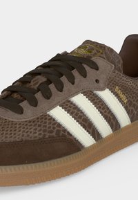Καφέ αθλητικό παπούτσι Adidas Samba με υφασμάτινο επάνω μέρος, λευκές ρίγες, μαύρα κορδόνια και λεπτομέρεια σόλας από καουτσούκ σε άσπρο φόντο.