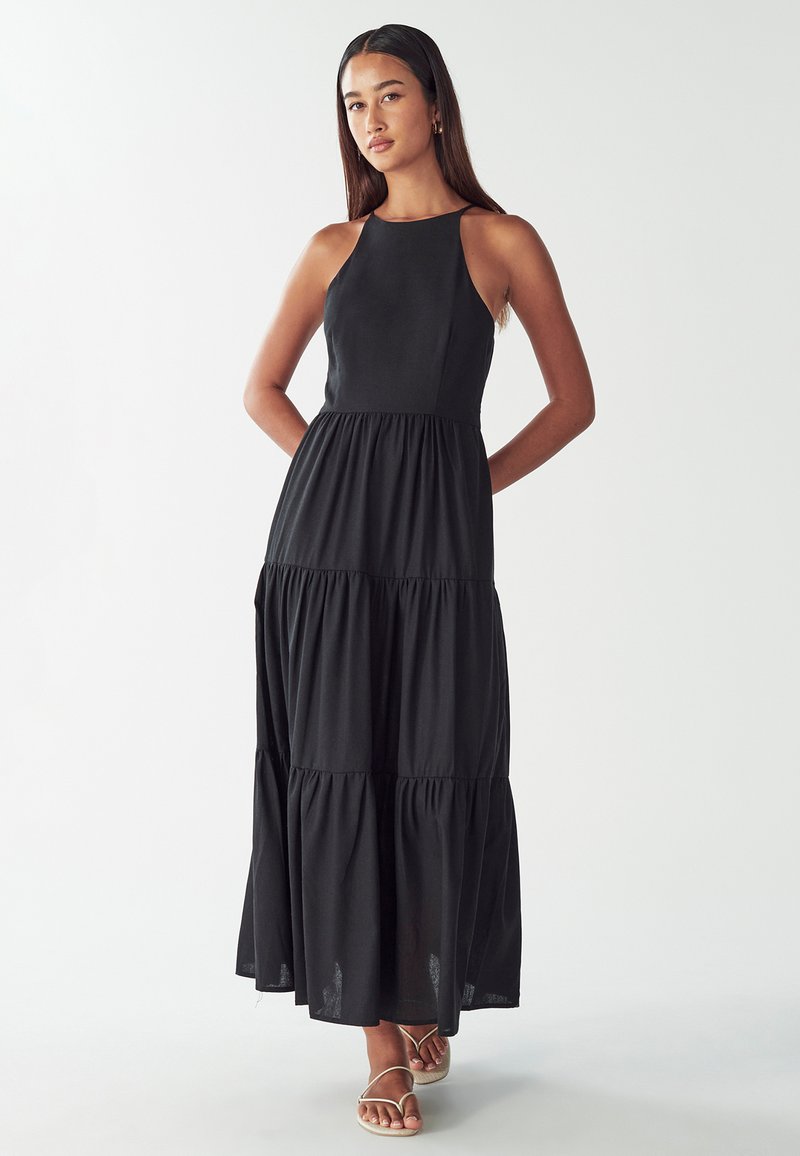 Musta maxi-mekko, jossa on istuva bodies, halterneck ja kerroksellinen hame. Valmistettu pehmeästä kankaasta, jonka pinta on sileä. Minimalistinen muotoilu.