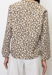 Blouse à manches longues en tissu léger, présentant un fond marron avec des motifs floraux crème, un col plissé et une coupe décontractée.