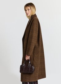 Brauner Karokemantel mit lockerer Passform, breitem Kragen und großen Knöpfen. Das Modell hält eine kleine dunkelbraune Handtasche mit silbernen Zubehör.