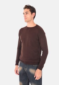 Maglione marrone lavorato a maglia, collo rotondo, polsini e orlo a coste, stile aderente, abbinato a jeans blu scoloriti, texture liscia.