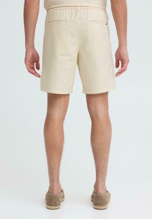 Mand iført beige shorts med baglommer og lysebrune slip-on sko, stående mod en ensartet lys baggrund.