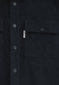 Ciemnoniebieska koszula z korduroy z pionowym prążkowanym wzorem, z dwoma kieszeniami na piersi, zapięciem na guziki oraz małą metką z boku.