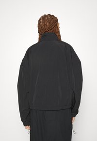 Czarna, lekka kurtka z wysokim kołnierzem, elastycznymi mankietami i luźnym krojem. Gładka faktura z delikatnym połyskiem, minimalistyczny design.