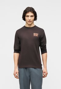 Νεαρός άνδρας με μεσαίου μήκους σκούρα μαλλιά, φορώντας μαύρο μακρυμάνικο πουκάμισο με πορτοκαλί γράμματα και μπλε παντελόνι, στέκεται μπροστά σε απλό φόντο.