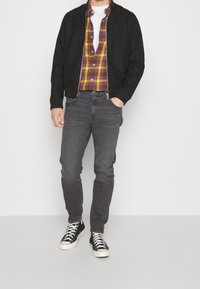 Black bomber jacket over a yellow and purple plaid shirt, paired with gray jeans and black high-top sneakers. Smooth, casual design.