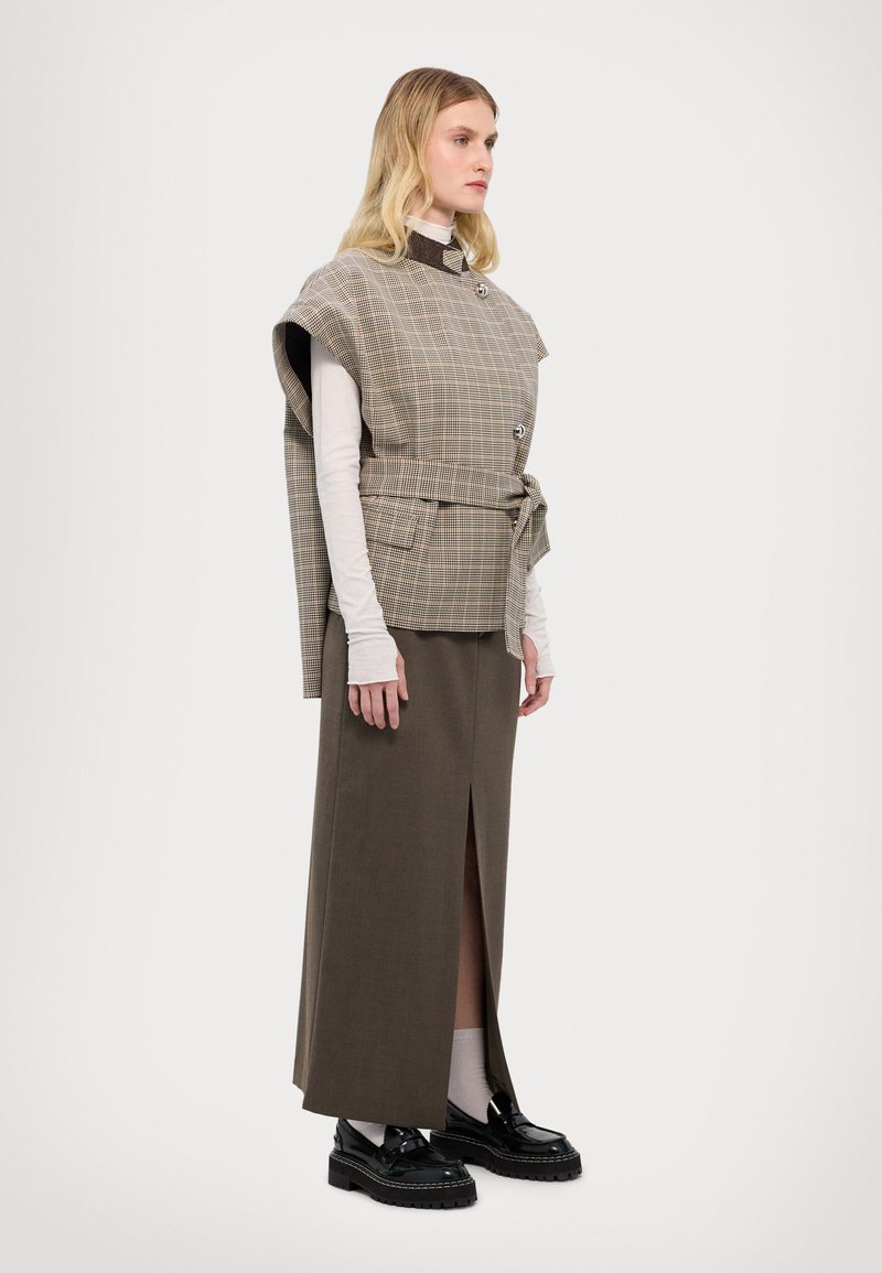Gilet a quadri beige con cintura e tasche laterali sopra una lunga gonna marrone con spacco frontale, abbinato a scarpe con plateau nere.