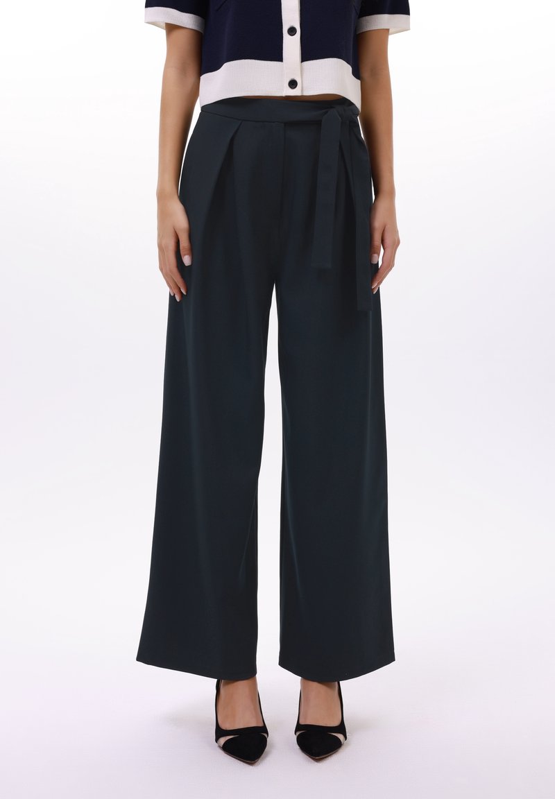Femme portant un pantalon noir taille haute à jambes larges avec une ceinture nouée, associé à des talons noirs pointus et un top court bleu marine et blanc boutonné.