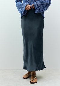 Langer, dunkelgrüner Satrock, kombiniert mit einem strukturierten blauen Pullover. Verfügt über eine glatte Oberfläche und een diskreten Glanz. Getragen mit braunen Sandalen.