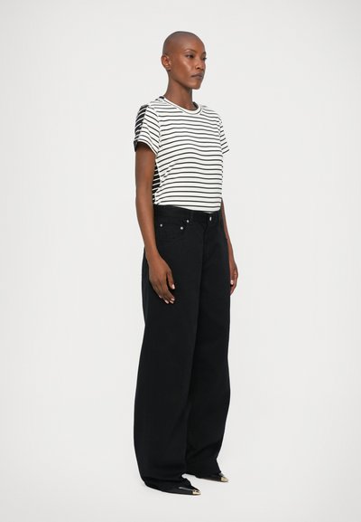 Gestreiftes Baumwoll-T-Shirt in Weiß und Schwarz, kombiniert mit weit geschnittenen schwarzen Jeans. Bemerkenswerte Seitendetails am Shirt. Model steht im Profil.