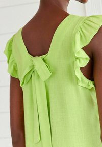 Top sans manches vert lime avec des bretelles volantes, un grand nœud à l'arrière et un tissu léger et texturé.