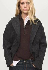 Donkergrijze wollen jas met een dubbelzijdig ontwerp, grote revers en twee voorzakken, gelaagd over een bruine gebreide trui en een witte blouse met kraag.
