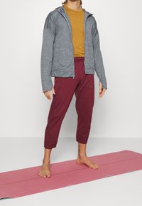 Grijze ritsjack met textuur stof, in combinatie met bordeauxrode cropped broek. Staande op een roze yogamat, op blote voeten, zonder zichtbare merkvermelding.