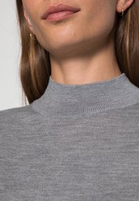 Gros plan sur le bas du visage et le cou d'une personne portant un pull à col montant côtelé gris et de petites boucles d'oreilles en or.