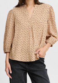 Blouse plissée beige avec un col en V, motifs micro noirs et manches trois-quarts. Le tissu a une finition texturée. Associée à un jean foncé.
