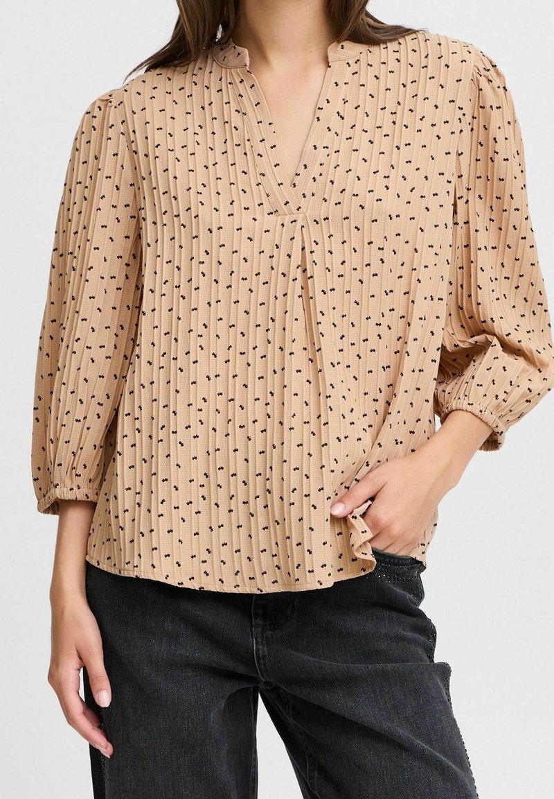 Blouse plissée beige avec un col en V, motifs micro noirs et manches trois-quarts. Le tissu a une finition texturée. Associée à un jean foncé.