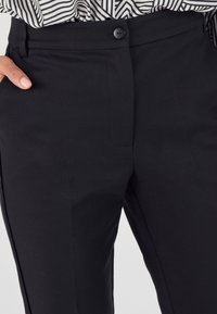Pantaloni neri su misura con una texture liscia, chiusura con bottone e tasche laterali. Il design include dettagli di pieghe per un aspetto strutturato.