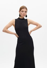 Vestido negro sin mangas de tejido suave, con un cuello alto adornado, bolsillos laterales y una silueta ajustada.