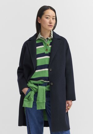 Frau trägt einen marineblauen Mantel über einem grün-navy gestreiften Hemd, mit einem grünen Pullover um die Taille gebunden und blauen Jeans.