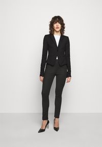 Blazer noir ajusté avec un bouton unique, associé à un pantalon foncé ajusté et des escarpins noirs à talons aiguilles, sur fond blanc uni.