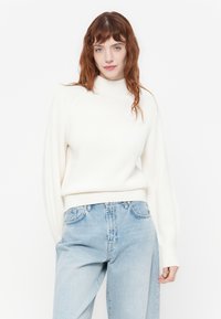 Hvit strikket genser med høy hals og ribbestruktur, sammen med lyseblå avslappede jeans. Søm detaljer på ermer.