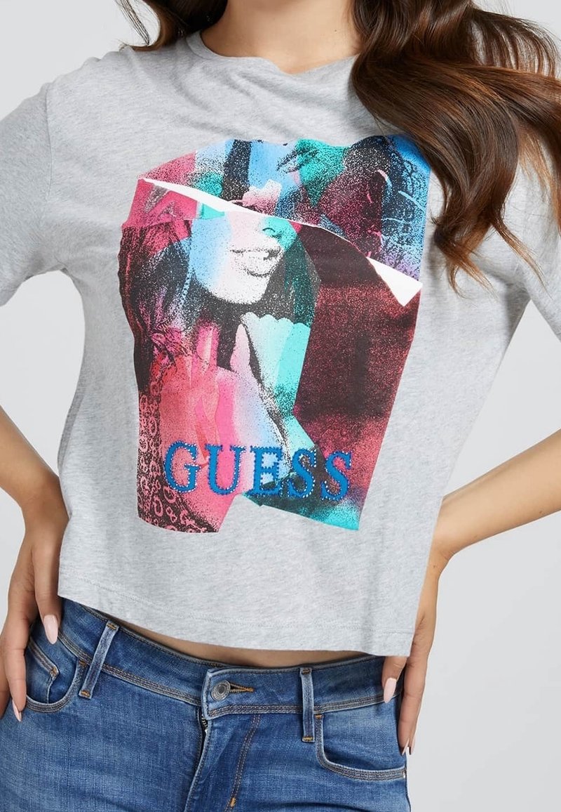 Šedé zkrácené tričko s barevným grafickým designem obsahujícím různé tvary a textury. Potisknuto nápisem "GUESS" modrými písmeny.