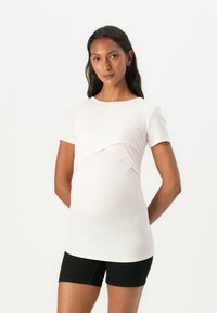 Top de maternitate ribbed roz deschis, cu detaliu răsucit în față, mâneci scurte și o croială lejeră, asortat cu pantaloni scurți negri.