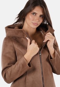 Giacca in finta pelle scamosciata marrone con cappuccio, chiusura con zip, maniche lunghe e una texture morbida. Modella che tiene il colletto, mostrando la sua finitura liscia.