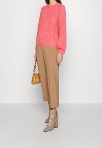 Rosa gestrickter Pullover mit langen Ärmeln, kombiniert mit hellbraunen cropped Hosen. Beigefarbene Absatzstiefeletten und eine kleine tanfarbene Schultertasche.