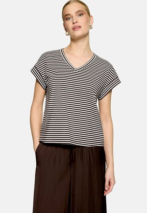 Femme portant un haut à manches courtes rayé noir et blanc avec un col en V, pantalon large marron foncé, mains dans les poches, sur un fond uni.