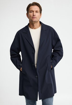 Homme portant un manteau bleu marine boutonné par-dessus une chemise beige, les mains dans les poches du manteau, debout devant un fond clair uni.